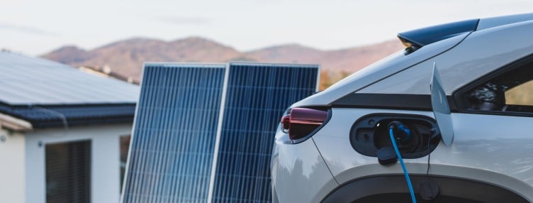 A silver-grey electric vehicle (EV) is charging in a scenic, desert destination in front of solar panels mounted on the ground and a white building with solar panels on the roof.