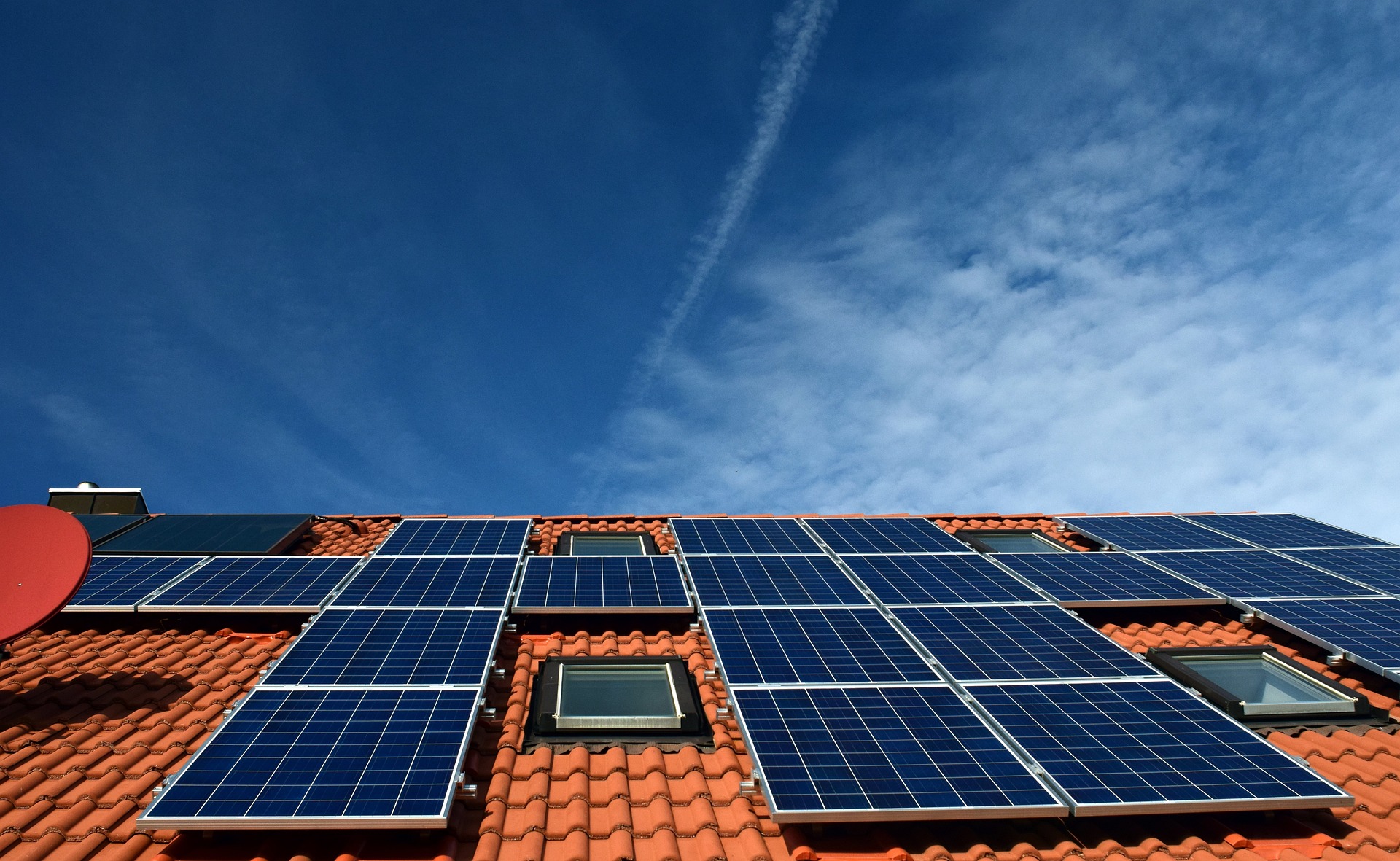 Residential solar panels on red tiled roof with partially cloudy blue sky in background.