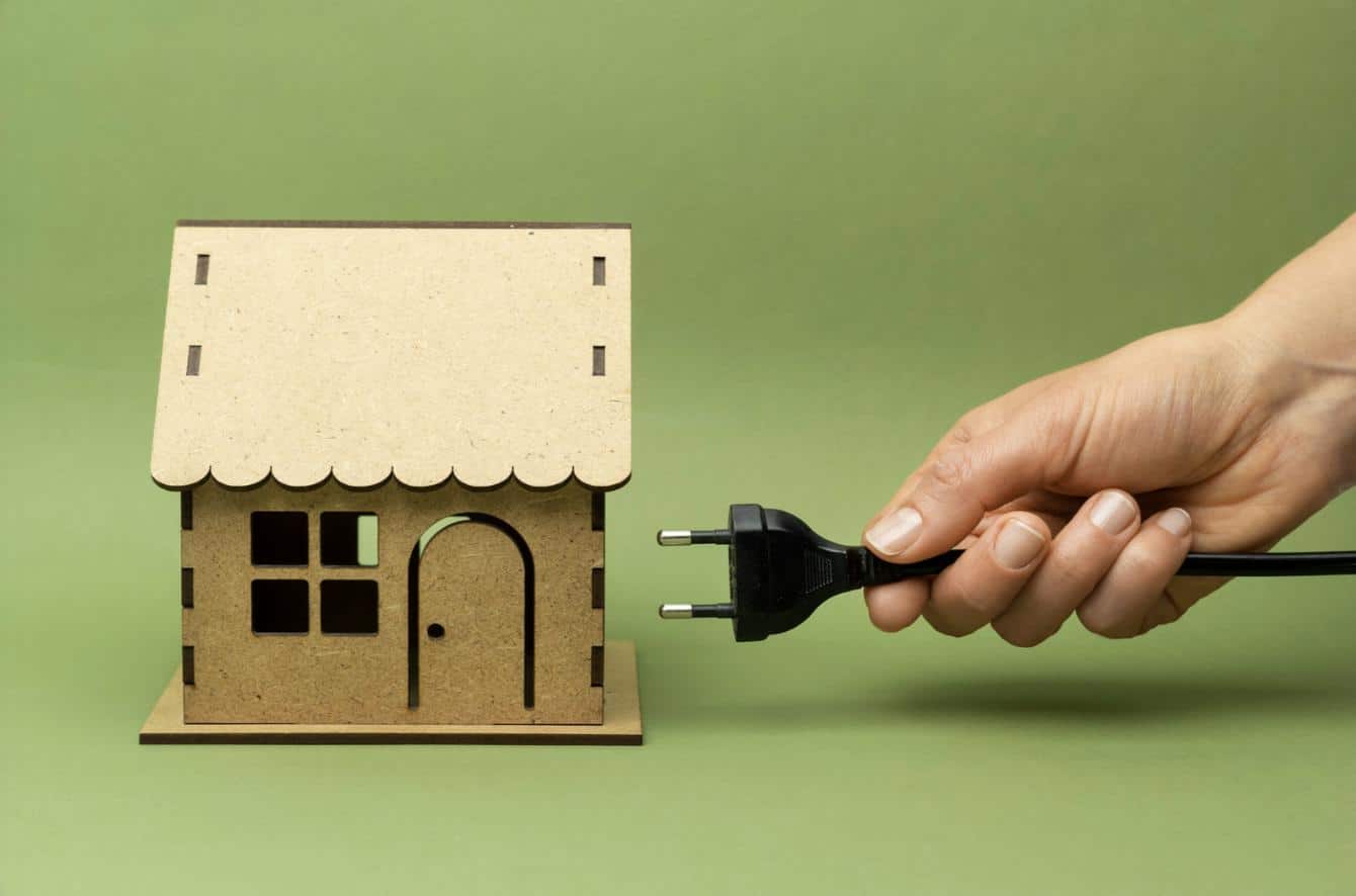 Hand holding power plug next to small wooden house model on green background, representing home electricity, solar energy, and sustainable power solutions.