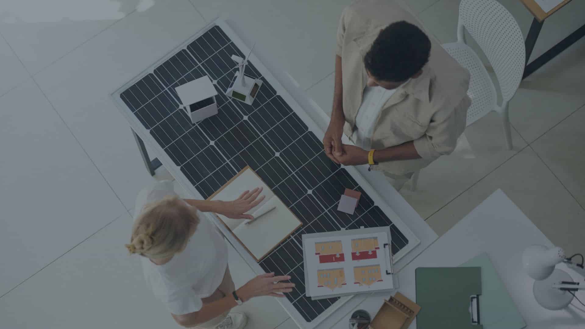 Two people reviewing solar project plans at a table with a large solar panel surface, miniature models, and design diagrams.